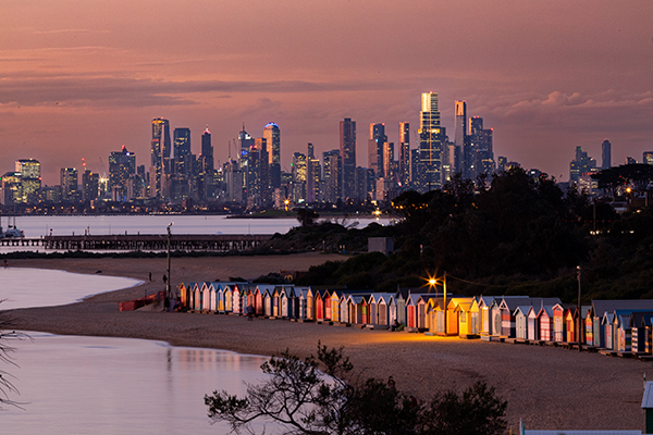 Brighton Beach Bathing Boxes lit by a street lamp and the glow of sunset at dusk with Melbourne CBD in the background