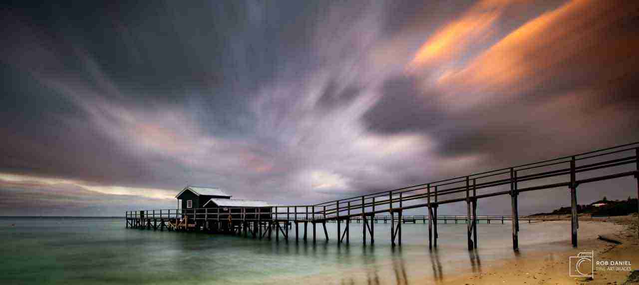 Photo of a jetty with a shack on it with rain clouds lit by the setting sun in the background